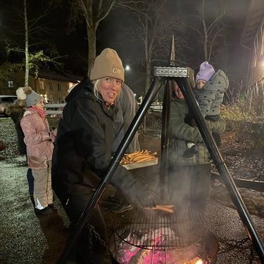 En grupp människor står utomhus i mörkret runt en eldstad med galler. En person i förgrunden bär keps och mörka kläder, medan två andra står bakom personen. Rök stiger från elden och området är upplyst av varmt ljus.