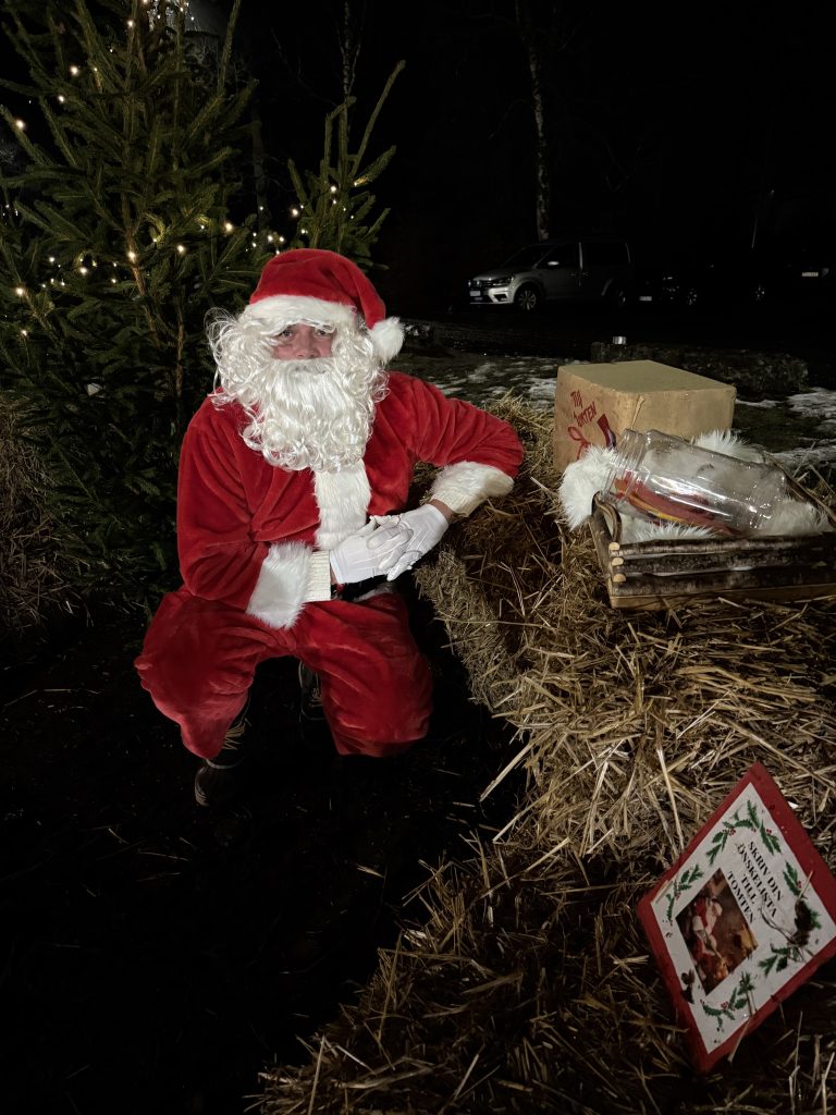 En man sitter utklädd till jultomte, bakom honom står en gran med en ljusslinga i sig.
