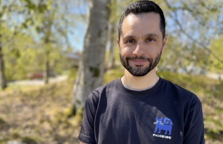 En man med mörkt, kort hår och skägg står utomhus framför björkar och grönska en solig dag. Han ler mot kameran och bär en mörk t-shirt med ett tryck av en blå katt och japansk text. Under bilden syns en bildtext om att brännboll väljs bort på vissa håll när elever känner press, men att idrottsläraren i bilden hittat ett annat sätt att lösa det.