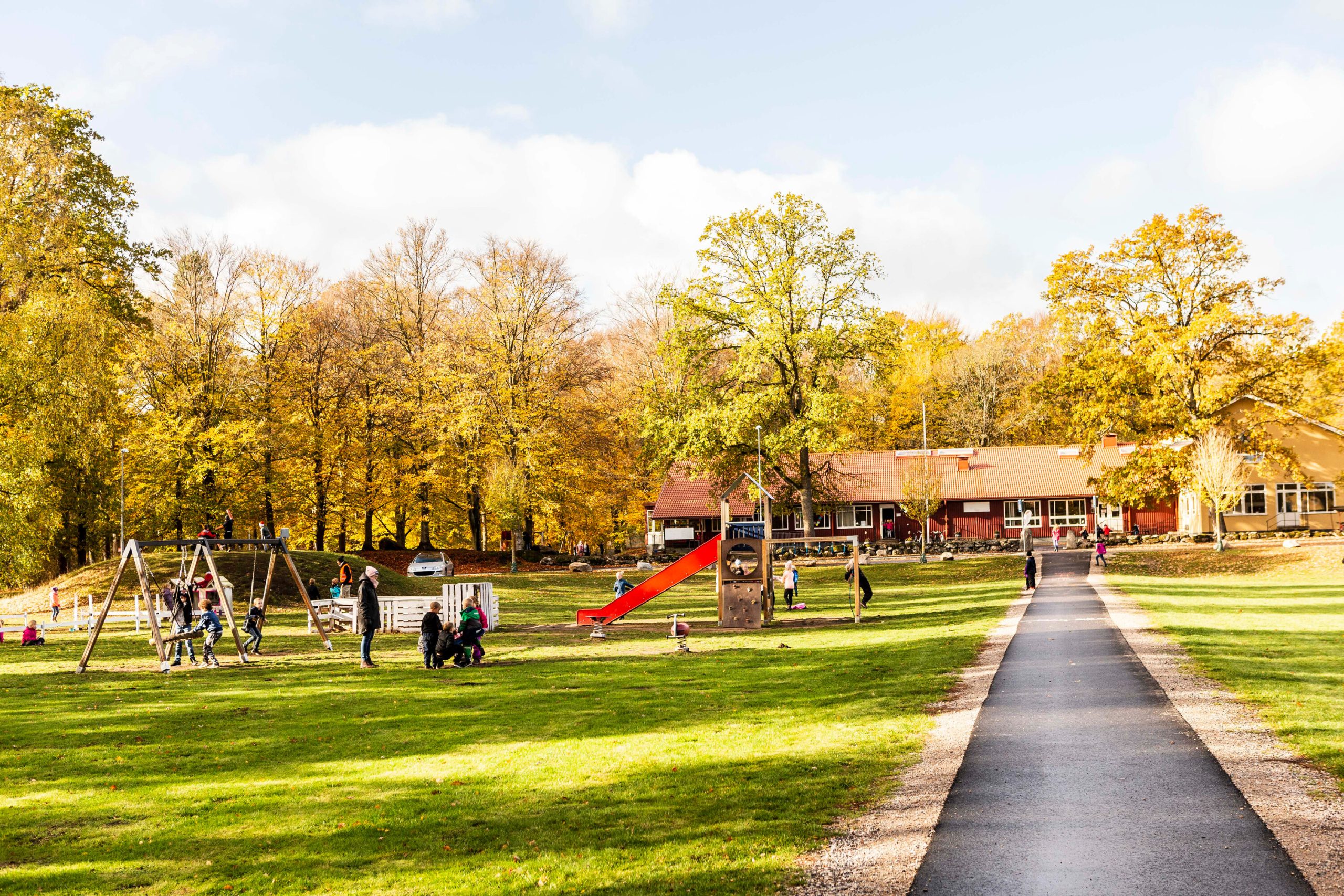 : Barn leker på en lekplats med gungor och rutschkana i en park med höstfärgade träd, med en röd skolbyggnad synlig i bakgrunden en solig höstdag.