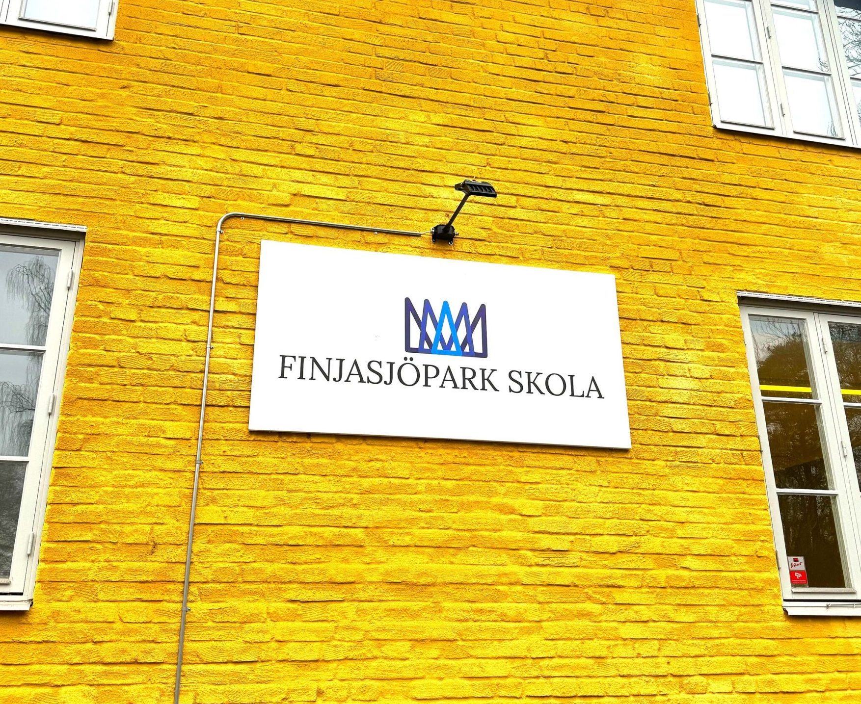 Fasaden på en gulmålad tegelbyggnad med vita fönster, med en vit skylt med skolans logotyp och texten "Finjasjöpark skola" fäst på väggen.