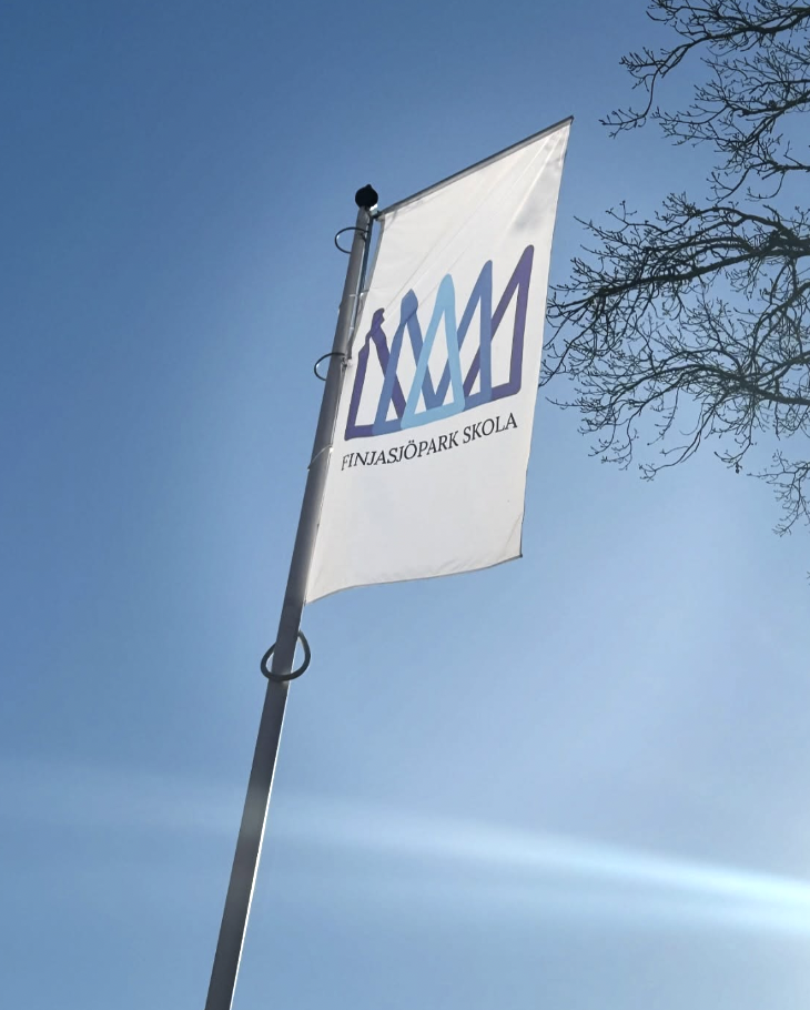 En vit flagga med Finjasjöpark skolas logotyp vajar på en flaggstång mot en klarblå himmel, med kala trädgrenar synliga i bildens övre hörn.