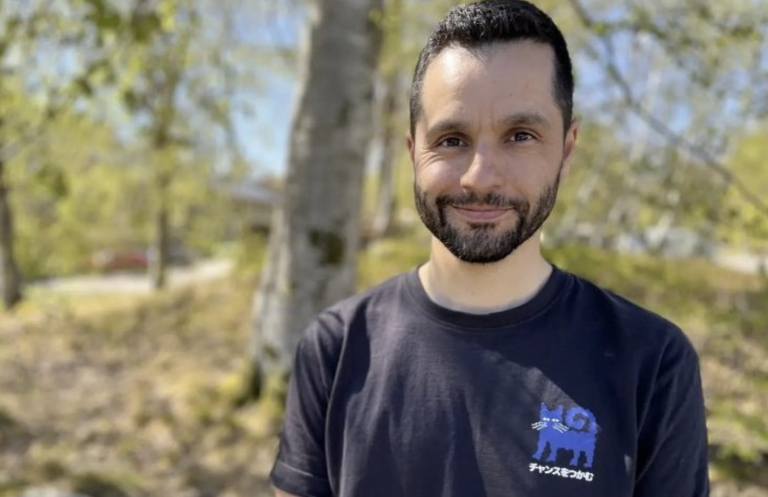 En man med mörkt, kort hår och skägg står utomhus framför björkar och grönska en solig dag. Han ler mot kameran och bär en mörk t-shirt med ett tryck av en blå katt och japansk text. Under bilden syns en bildtext om att brännboll väljs bort på vissa håll när elever känner press, men att idrottsläraren i bilden hittat ett annat sätt att lösa det.